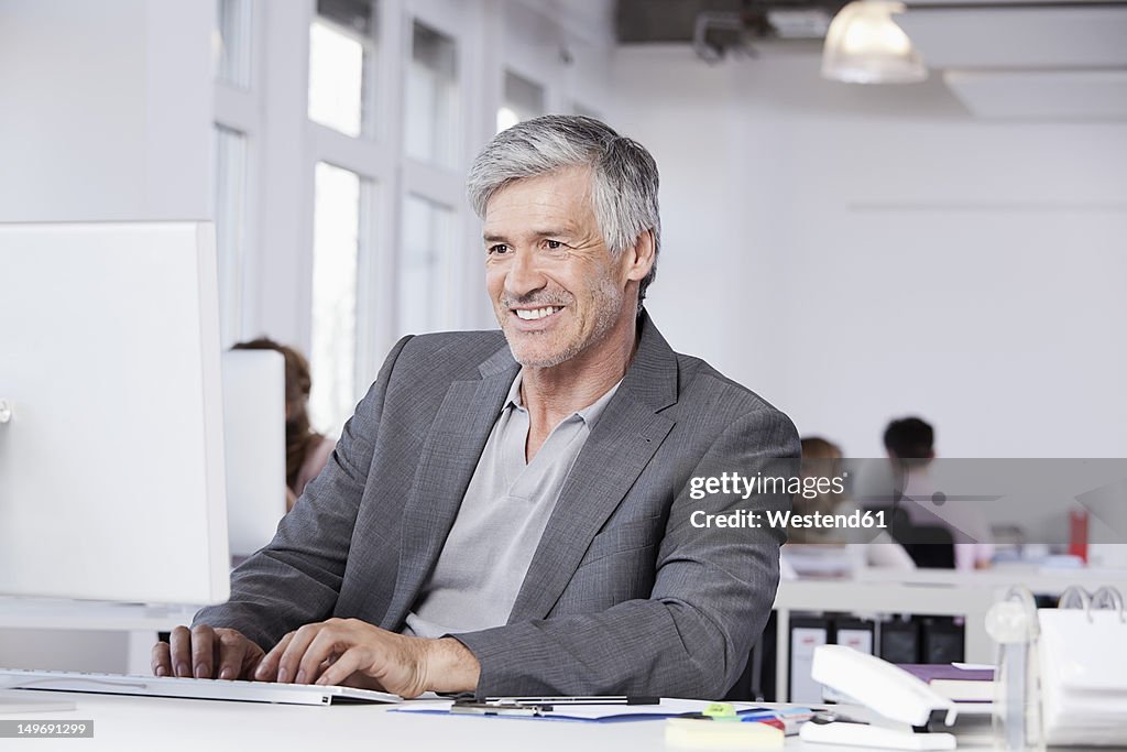 Germany, Bavaria, Munich, Mature man using computer, colleagues working in background