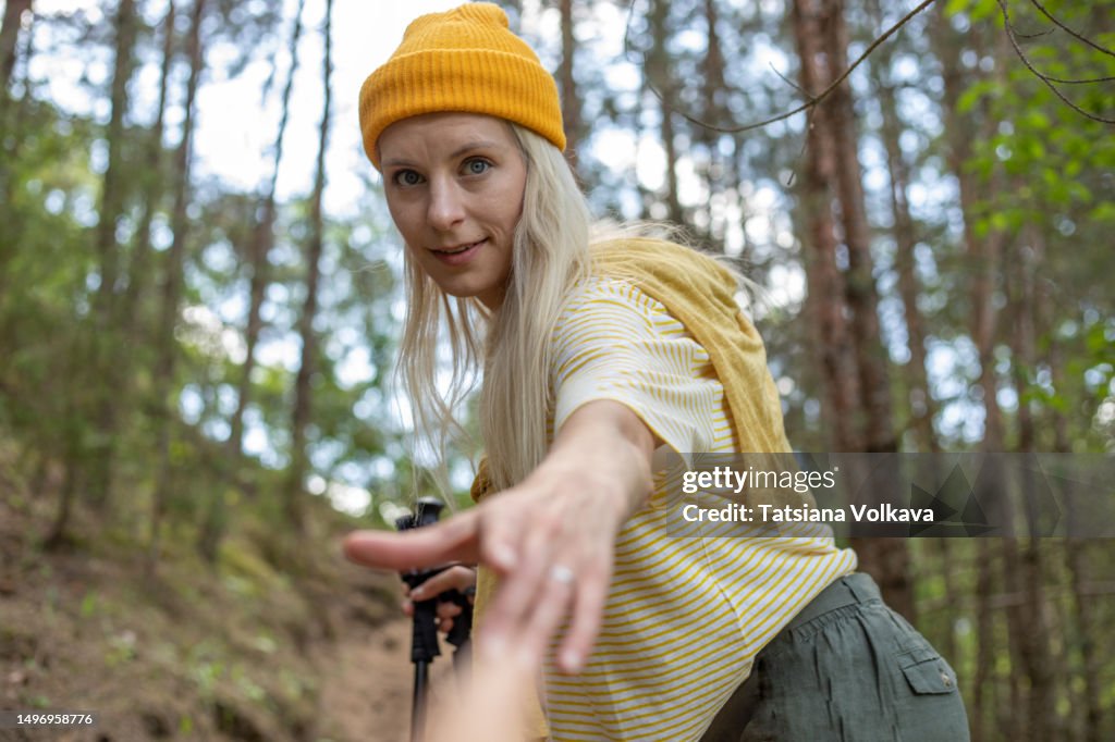A blonde woman in a yellow hat, engaged in Nordic walking, was elected to the mountain, turned around and extends a helping hand to the person following her.