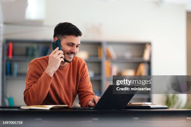 smiling businessman talking on a mobile phone while working on his laptop - friendly business phonecall stock pictures, royalty-free photos & images