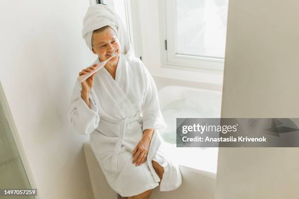 senior woman brushing teeth with electric toothbrush in bathroom. - brush teeth phone stock pictures, royalty-free photos & images