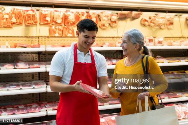 latin american friendly salesman suggesting a fresh product to senior female customer at the refrigerated meat section of the supermarket - latin american and hispanic shopping bags stockfoto's en -beelden
