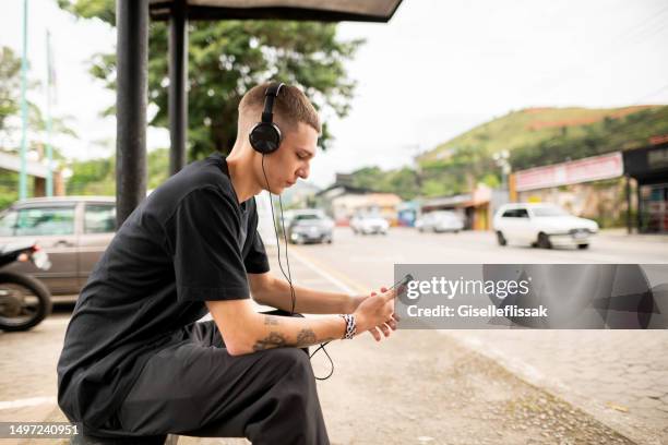 junger mann, der mit kopfhörern an der bushaltestelle wartet und auf sein handy schaut - bus bench stock-fotos und bilder