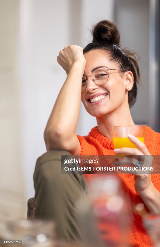 Portrait of a beautiful mature woman with hair bun and red tanktop sweater is looking at camera while having a glass of orange juice.