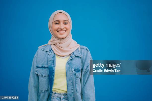 portrait asian malay woman with hijab looking at camera smiling standing in front of blue wall - hoofddoek stockfoto's en -beelden