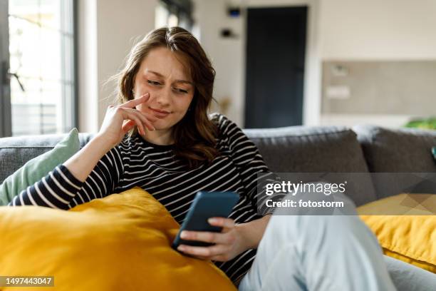 young woman lounging on the sofa and scrolling online via smart phone - scrollen stockfoto's en -beelden