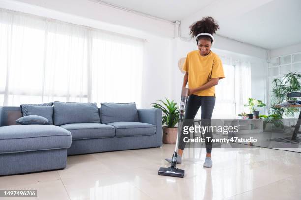 a black young woman listening to music while using a vacuum cleaner cleaning the room - vacuum cleaner woman stockfoto's en -beelden