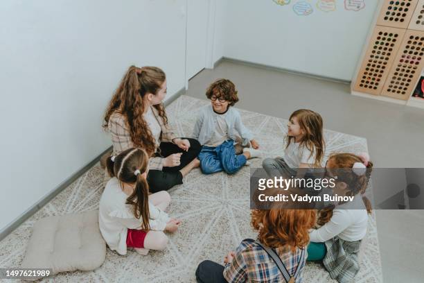 schoolchildren with their teacher sitting on the floor in circle in a classroom - bambini seduti in cerchio foto e immagini stock