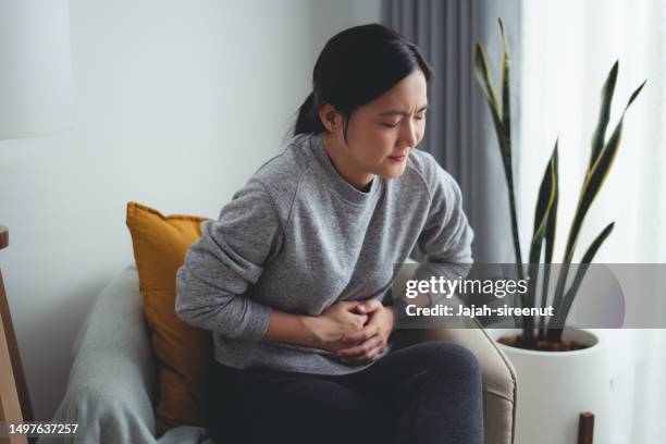 asian woman feeling stomachache sitting on armchair in living room at home. - mid adult women stock pictures, royalty-free photos & images