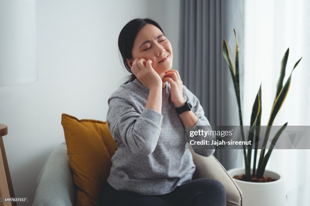 Asian woman irritate itching dry skin scratching her skin sitting on armchair in living room at home.
