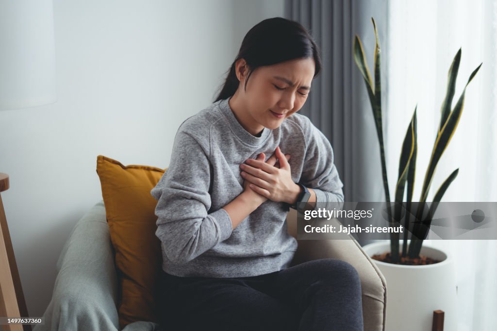 Asiatische Frau, die unter Brustschmerzen leidet, sitzt zu Hause auf einem Sessel im Wohnzimmer.