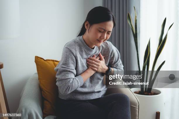 asiatische frau, die unter brustschmerzen leidet, sitzt zu hause auf einem sessel im wohnzimmer. - herzerkrankung stock-fotos und bilder