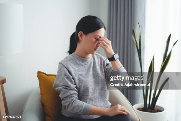 asian woman suffering from eyes pain sitting on armchair in living room at home. - rubbing eyes stock pictures, royalty-free photos & images