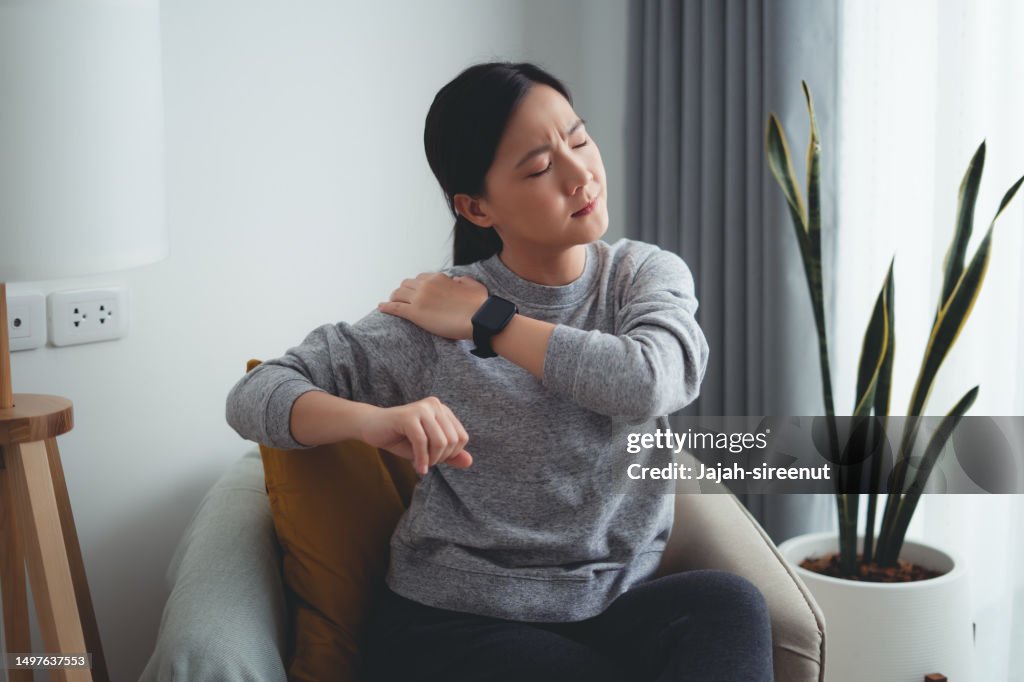 Asian woman suffering from shoulder pain sitting on armchair in living room at home.