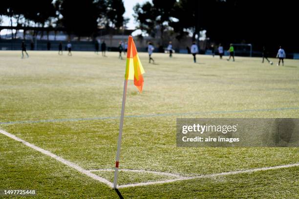 a yellow and orange corner flag of a soccer field covered with artificial turf with players in the background. - corner stock pictures, royalty-free photos & images
