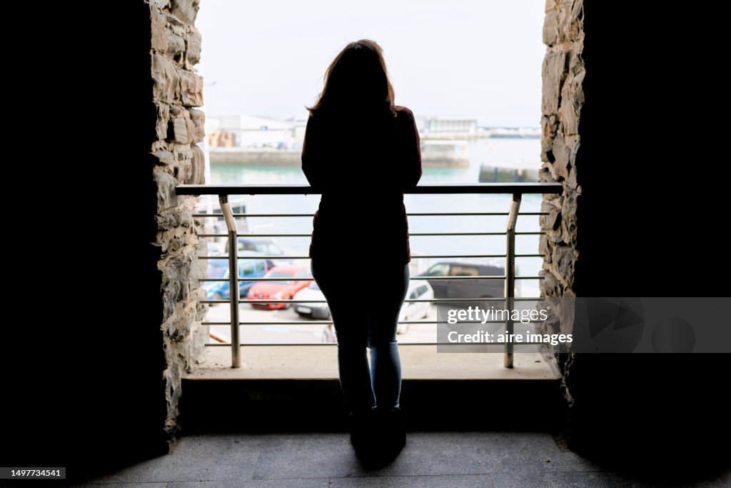 A silhouette of a woman with her back against the light leaning against a railing with columns of rocks, vehicles and the sea in the background.