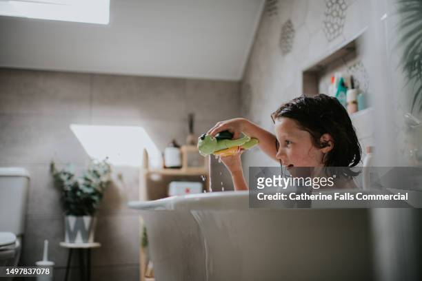 a child, with soapy hair, and a plastic bath toy, enjoys a warm bath. - free standing bath stock pictures, royalty-free photos & images