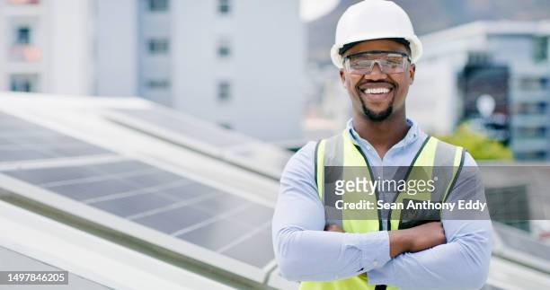ein junger afroamerikaner trägt eine brille und einen schutzhelm, während er ein solarpanel installiert. porträt eines selbstbewussten männlichen mitarbeiters, der auf einem gebäude auf einer baustelle steht - male looking content stock-fotos und bilder