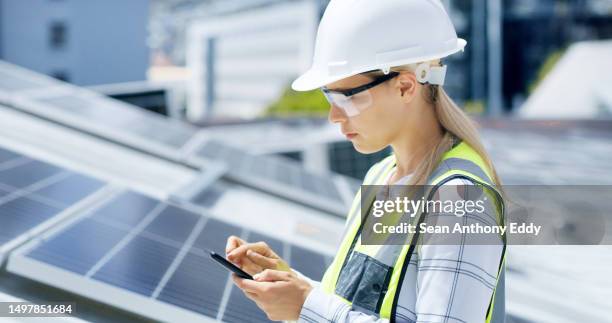 a serious young woman using a smartphone while installing solar panels on the roof of a building. a focused construction worker working online with her phone on a renewable energy project - female likeness stock pictures, royalty-free photos & images