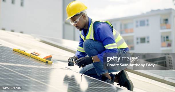 a young man using a drill, measuring tape and spirit level while installing solar panels on the roof of a building. a man working with tools on a renewable energy project - black roof stock pictures, royalty-free photos & images