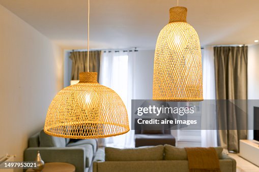 Two lamps hanging from the ceiling woven in yellow thread, lit at different levels in the background a living room with several furniture and cushions, with curtains and wall in white color