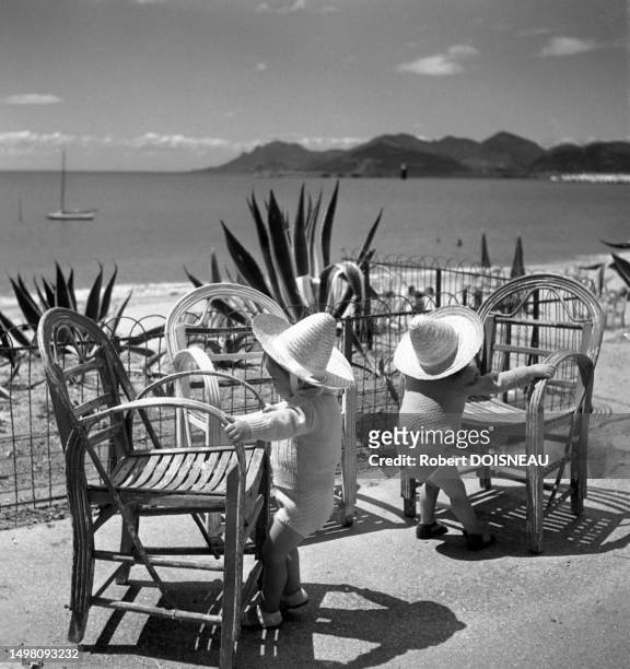 Deux enfants en bas âge avec des chapeaux de paille se tiennent debout chacun à une chaise en bois sur une terrasse face à la mer sur la Côte d'azur...