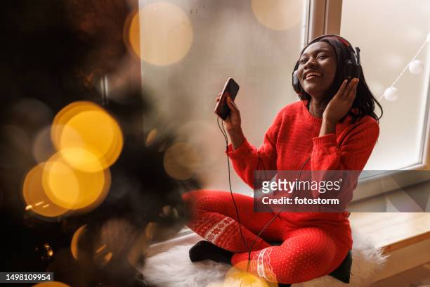 young woman listening to music via headphones on the window sill - christmas music listen stock pictures, royalty-free photos & images