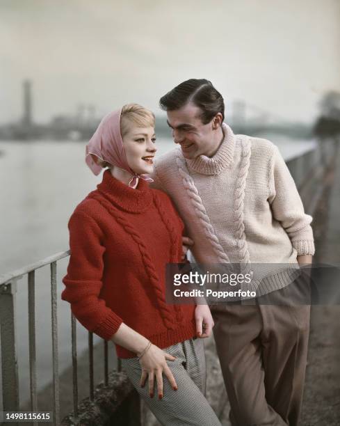 Posed exterior scene of two fashion models, one male and one female, wearing matching cable knit sweaters with roll necks in red and cream wool, they...
