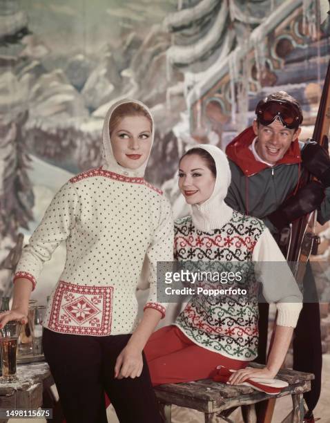 Posed studio portrait of two female fashion models and one male model standing in front of a ski scene backdrop, the women wear hooded knitted ski...