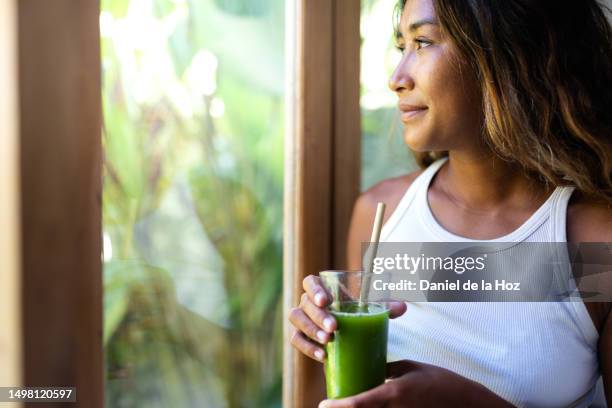 close up of happy young asian filipino woman looking at nature through the window holding glass of healthy green smoothie. copy space. healthy lifestyle and wellness - smoothie close up textfreiraum stock-fotos und bilder