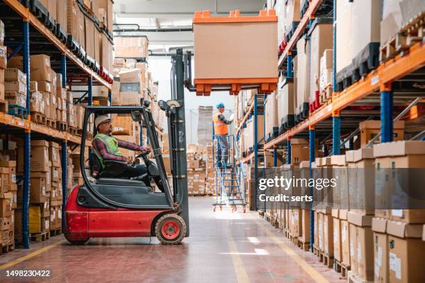 trabajador uniformado conduciendo y cargando cajas de cartón con cargador apilador de carretilla elevadora en un almacén de fábrica - almacén fotografías e imágenes de stock