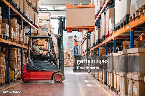 Uniformed Worker Driving and Loading Cardboard Boxes with Forklift Stacker Loader in a Factory Warehouse Uniformed Worker Driving and Loading Cardboard Boxes with Forklift Stacker Loader in a Factory Warehouse