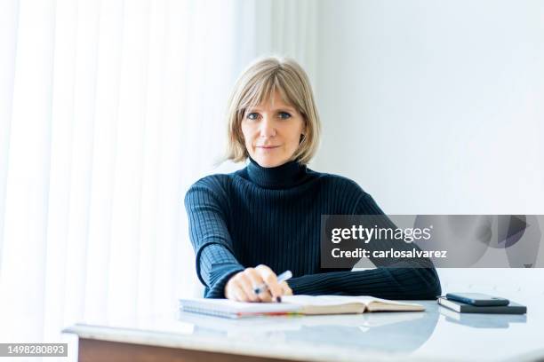 portrait of an adult blonde woman working at home - 50s woman writing at table stock pictures, royalty-free photos & images