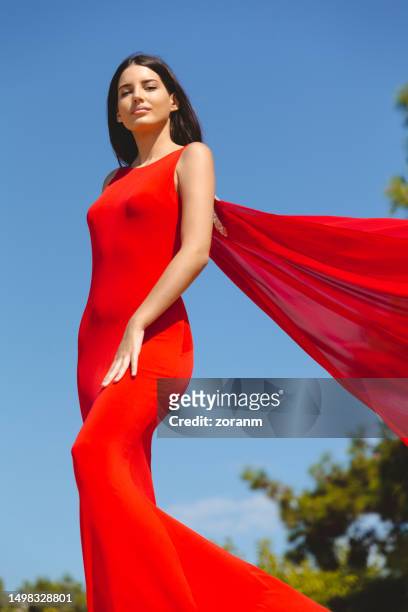 beautiful young brunette in long red sleeveless dress with train, standing outdoors - woman long dress beach stock pictures, royalty-free photos & images