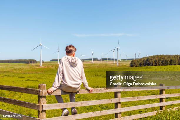 rear view of a boy sitting on a fence and looking ahead of a wind turbines farm in a countryside - ahead stock pictures, royalty-free photos & images