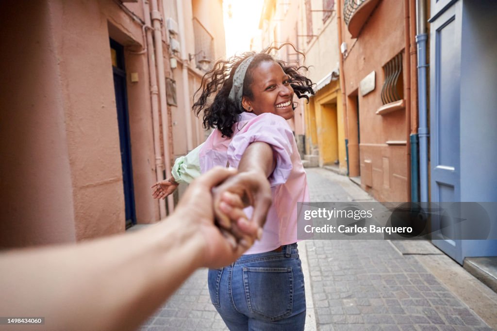 Happy woman running into a village alley pulling her boyfriend's hand and looking at him smiling.