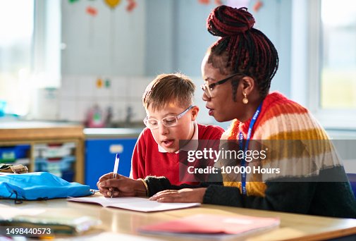 Black mid adult primary school teacher helping boy with Down syndrome in classroom Black mid adult primary school teacher helping boy with Down syndrome in classroom