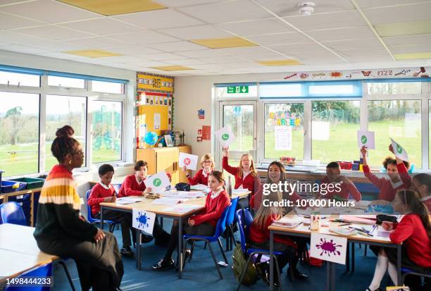 teacher talking to class of primary school children holding up pieces of paper - niveau primaire photos et images de collection