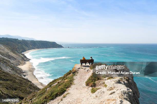 landscape of the asturian coast, view of the sea and rocky cliffs on which there is a bench on which two people are sitting with an unbeatable view on a blue sky day. - penhasco caraterísticas do território imagens e fotografias de stock