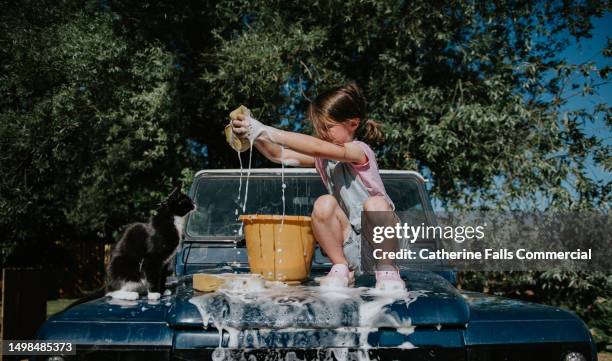 a little girl crouches on the bonnet of a car and washes it with a bucket and sponge. her pet cat watches. - lavagem automática imagens e fotografias de stock