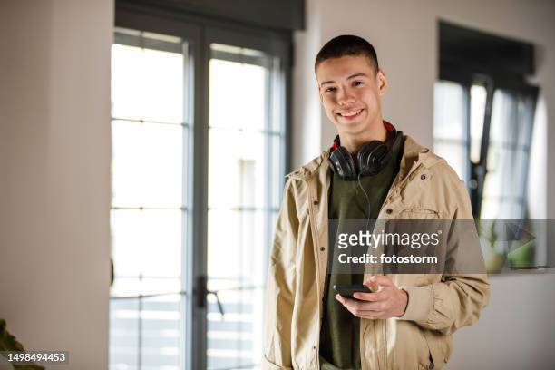 charming teenage boy standing in a room, holding smart phone, smiling with confidence - alleen tienerjongens stockfoto's en -beelden