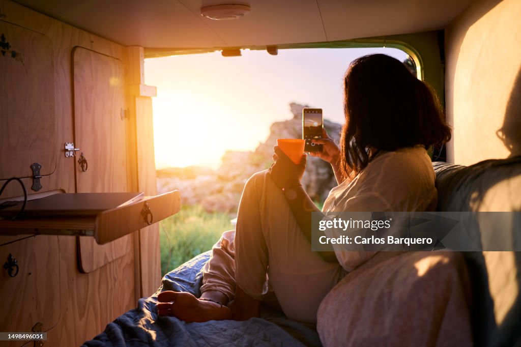 Unrecognizable woman enjoying the sunset inside a camper van, taking a photo for social media.