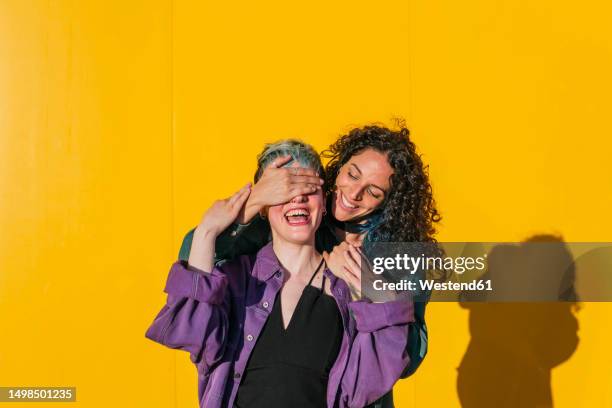 happy woman covering eyes of lesbian friend in front of yellow wall - hands covering eyes stock pictures, royalty-free photos & images