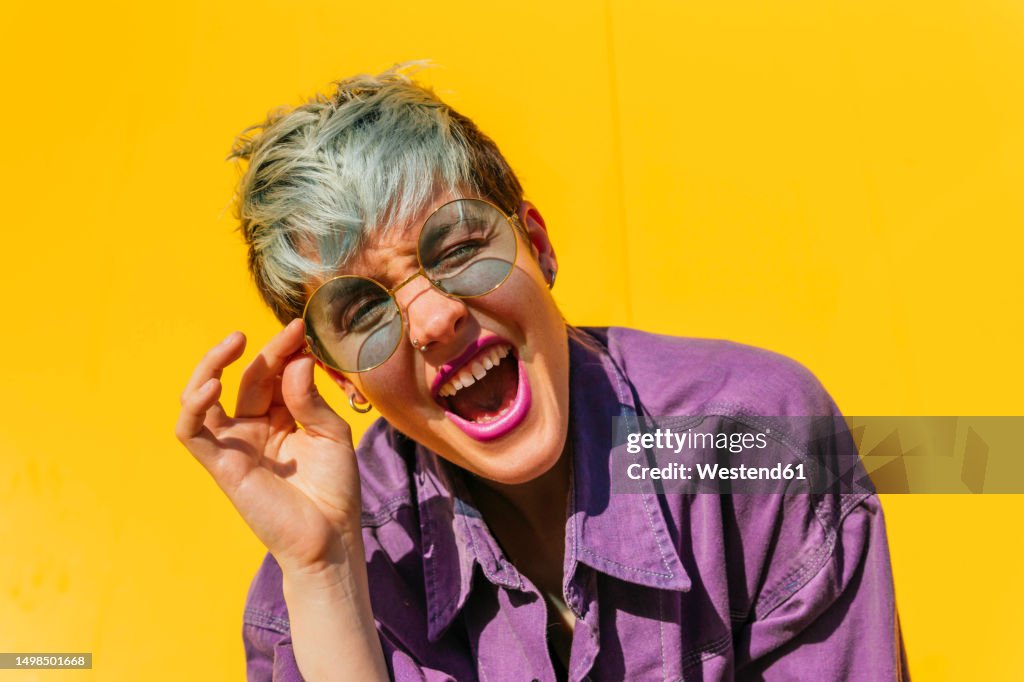 Carefree woman holding sunglasses and screaming in front of yellow wall