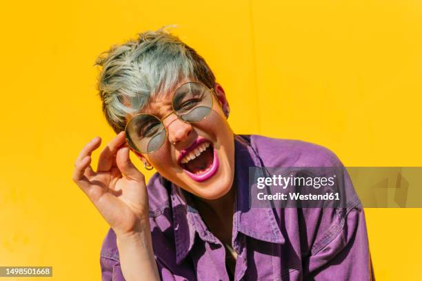 carefree woman holding sunglasses and screaming in front of yellow wall - purple jacket stock pictures, royalty-free photos & images