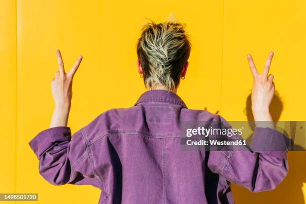 woman making peace sign facing yellow wall - denim jacket from behind stock pictures, royalty-free photos & images