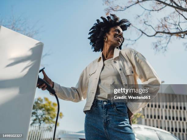smiling young woman charging electric car - stromtankstelle stock-fotos und bilder