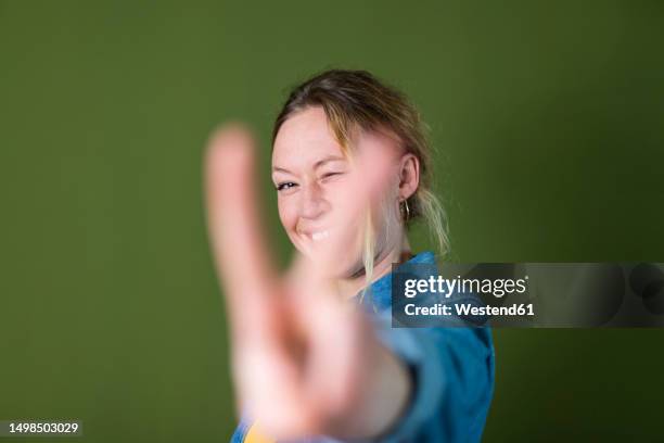 smiling young woman gesturing peace sign in front of green wall - victory sign stock-fotos und bilder