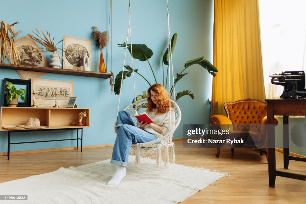 The beautiful young red-hair woman sitting on the hammock rope swing chair and reading a book in bright room