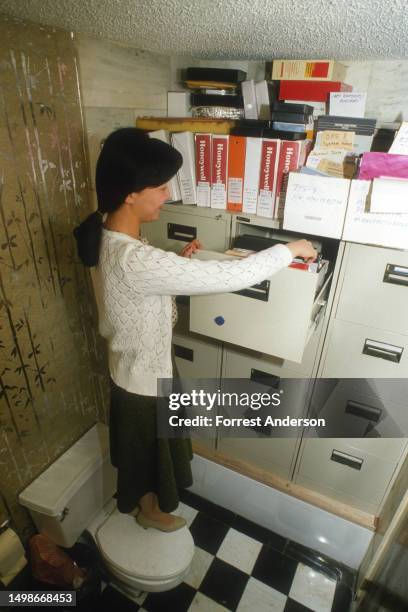 Elevated view of a woman as she stands on a toilet to look in a filing cabinet stacked in a bathtub in room at the Jianguo Hotel, Beijing, China,...