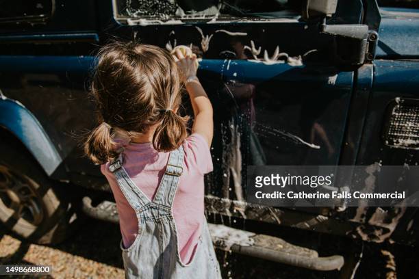 a little girl uses a yellow sponge to wash a blue car, the soap creates a lather that runs down the side of the car - hubcap stock pictures, royalty-free photos & images